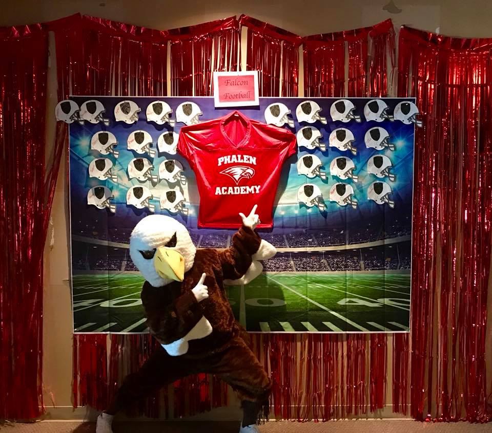 Phalen Falcon Mascot standing in front of bulletin football board. 